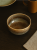 Okrągła ceramiczna miska z zacieranym, brązowo-beżowym szkliwem stoi na ciemnym drewnianym stole. Przytulna aranżacja obejmuje lnianą serwetkę, ochrowy kubek oraz talerz częściowo widoczny w tle.
